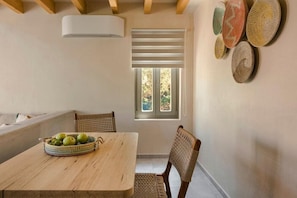 A dining area with a wooden table, wicker chairs, a basket of green apples, a window with blinds, and woven wall decorations. - Chromata Assos Villas Kefalonia (Kefalonia)