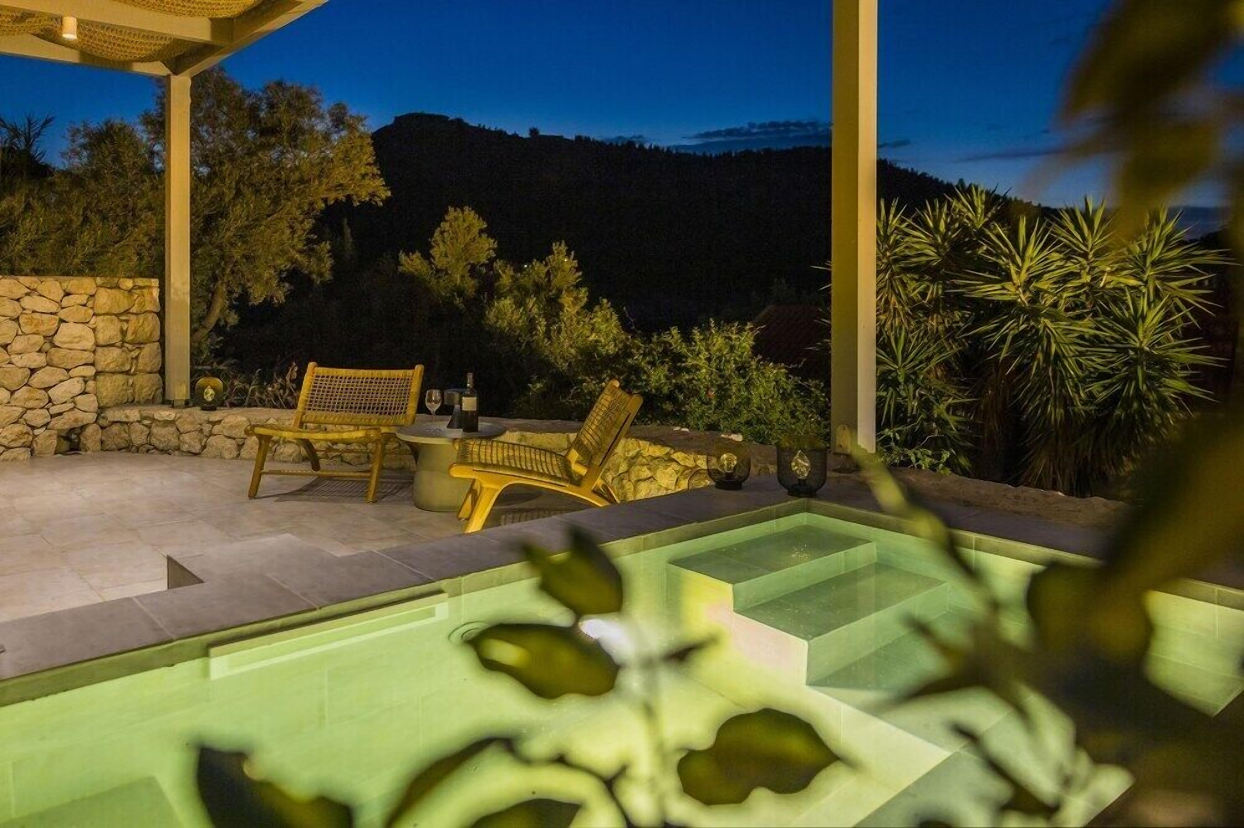 Zona de piscina con muro de piedra, dos sillas de mimbre y vista a una montaña al atardecer.