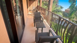 A balcony with a seating area, including a table and chairs, and a view of a garden with palm trees. - Ko Olina | Disney Family Adventure | Lagoon + Pool | 3BR Sleeps 12 (Kapolei)