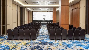 Un salón de conferencias con filas de sillas orientadas hacia una pantalla para presentaciones, suelo alfombrado y un diseño de techo moderno.