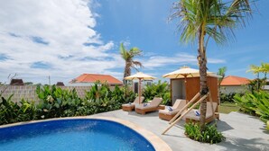 Una piscina con sillones de descanso y sombrillas, rodeada de vegetación y edificios con tejados rojos.