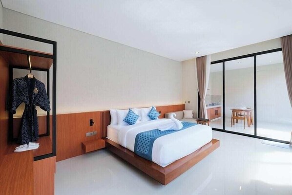 A modern hotel room with a large bed, a wooden nightstand, a mirror, and a view of the outside through a sliding glass door.