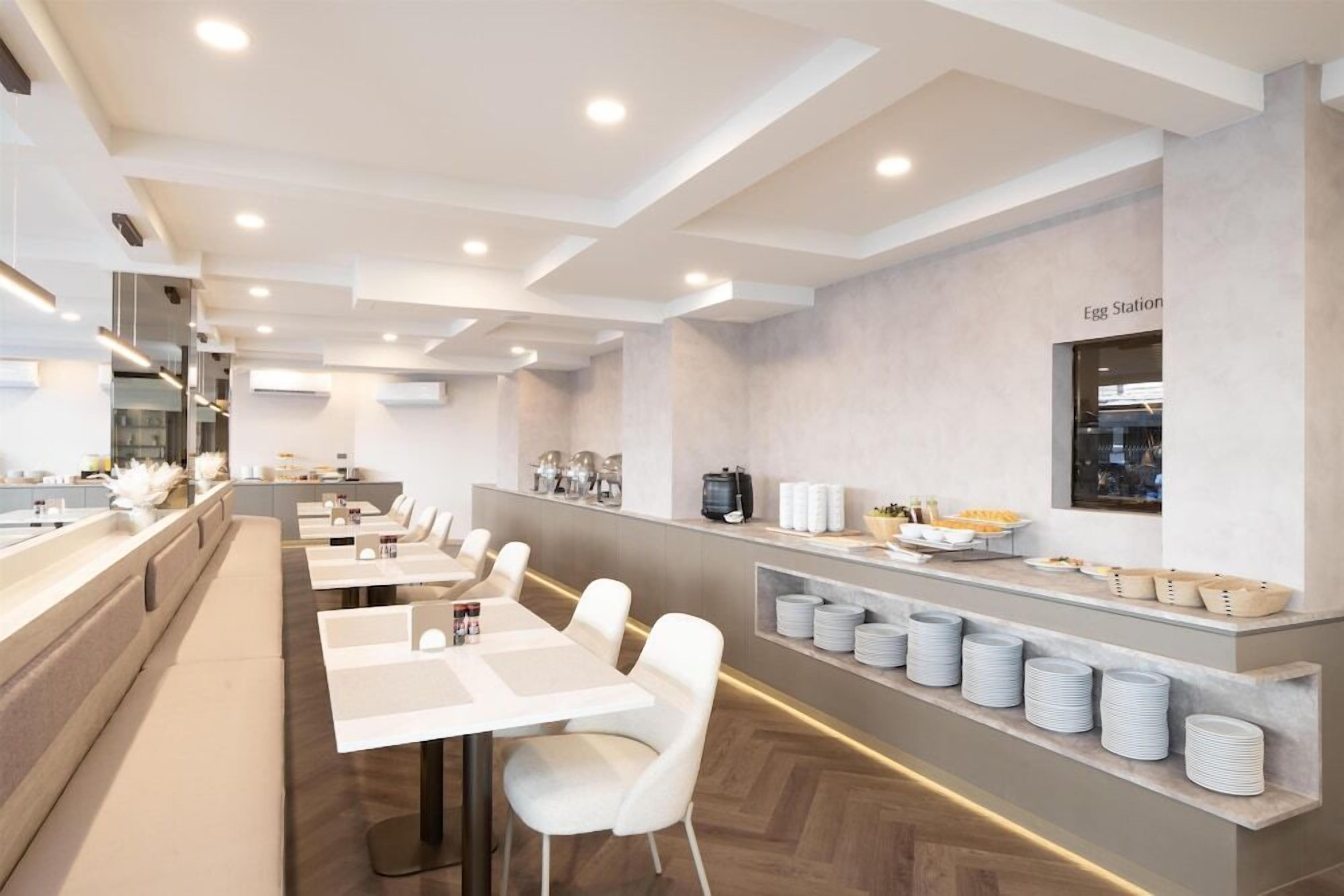A modern dining area with white tables and chairs, a counter with food items, and a wall-mounted sign.