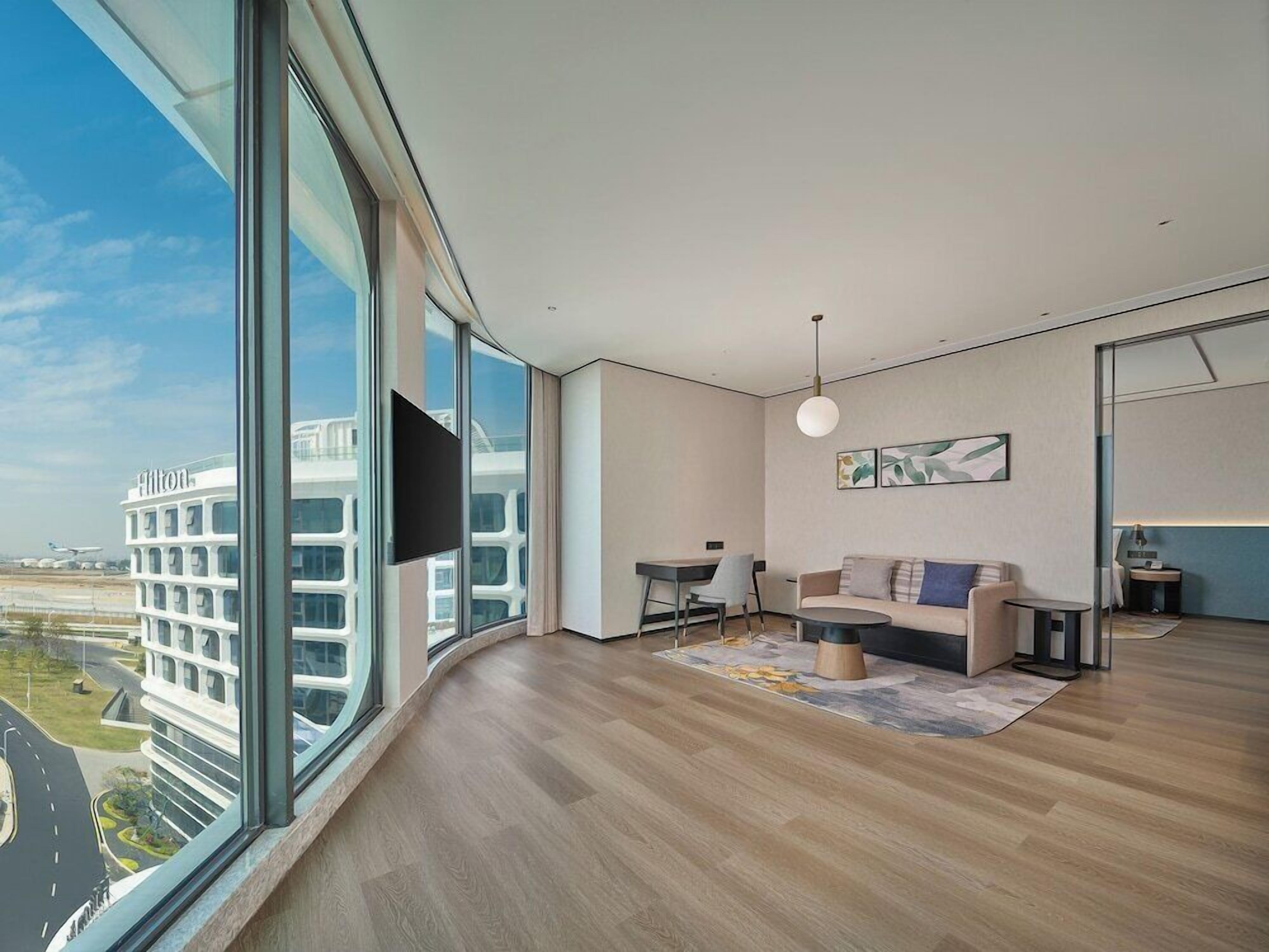 A modern hotel room with a large window, a flat-screen TV, a sofa, a coffee table, and a small side table.