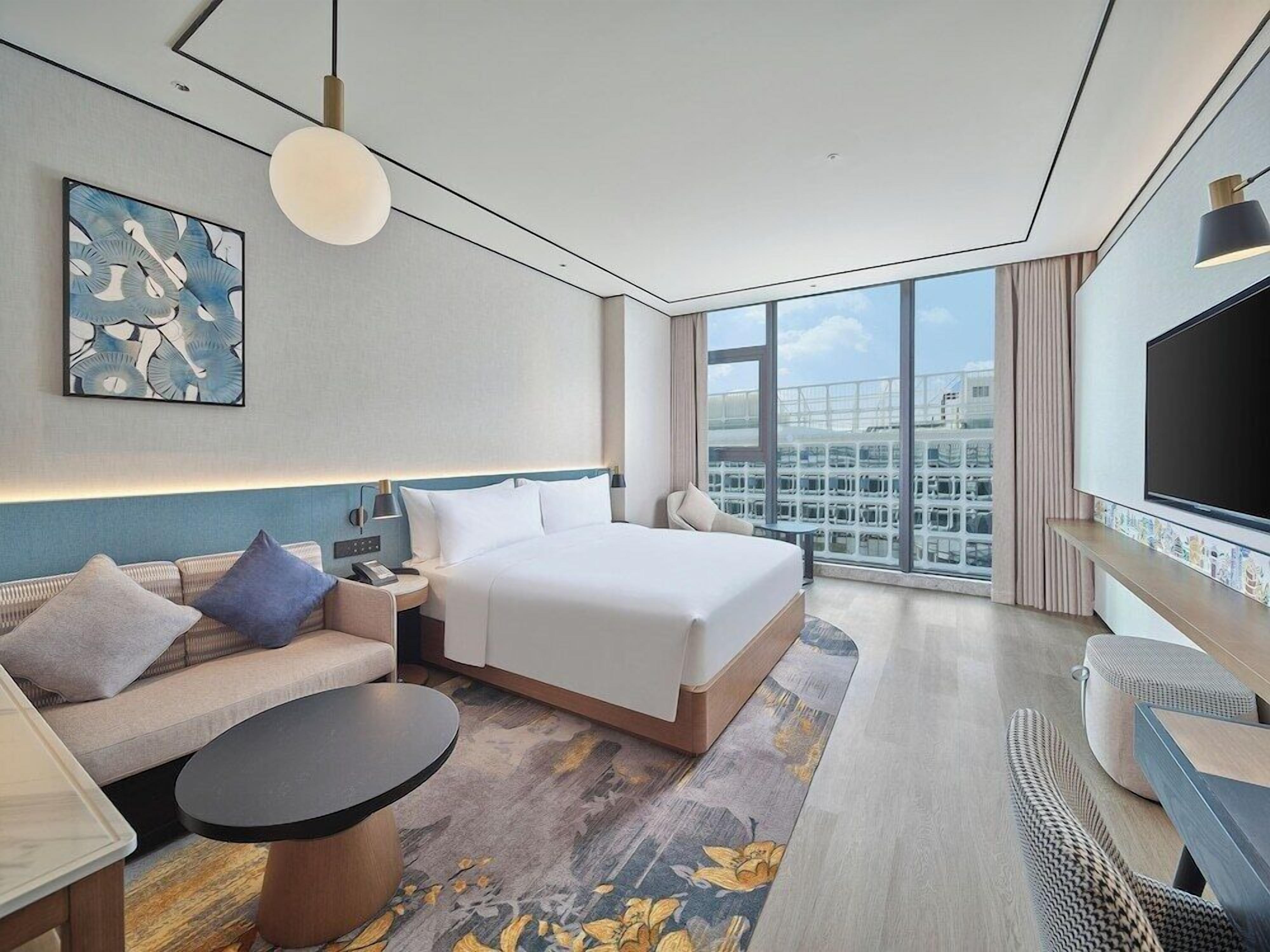 A modern hotel room with a large bed, a sofa, a small table, a TV, and a city view.