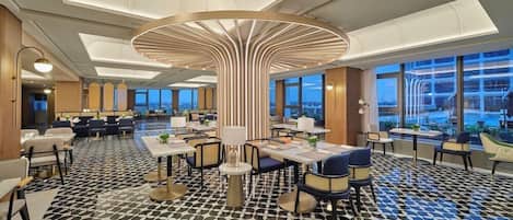 A modern hotel lobby with a central circular light fixture, dining tables, and chairs arranged for guests.