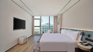 A modern hotel room with a large bed, a flat-screen TV, a bedside table with a lamp, and a view of the city through large windows.