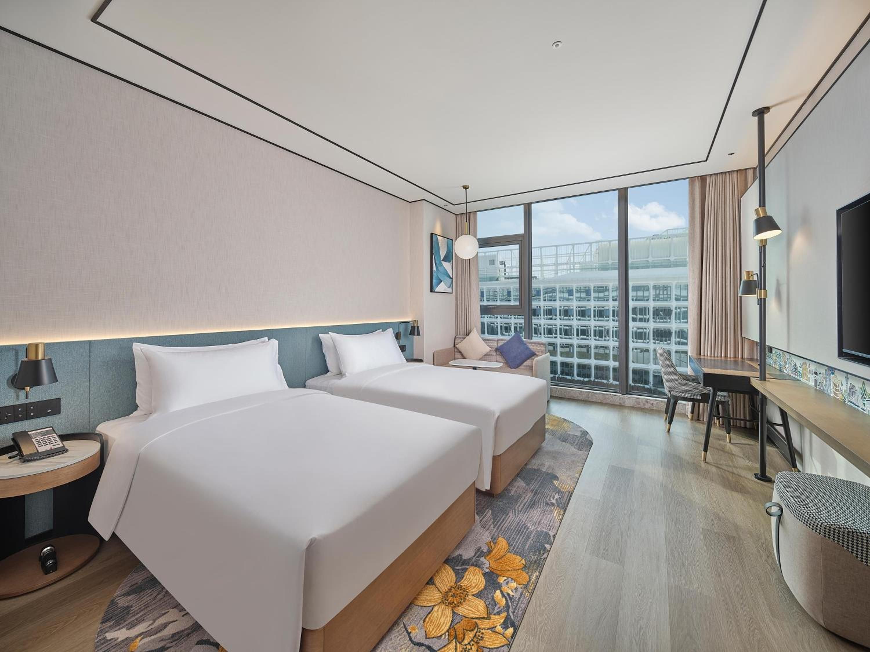 A hotel room with two beds, a desk, a chair, a TV, and a large window with a city view.