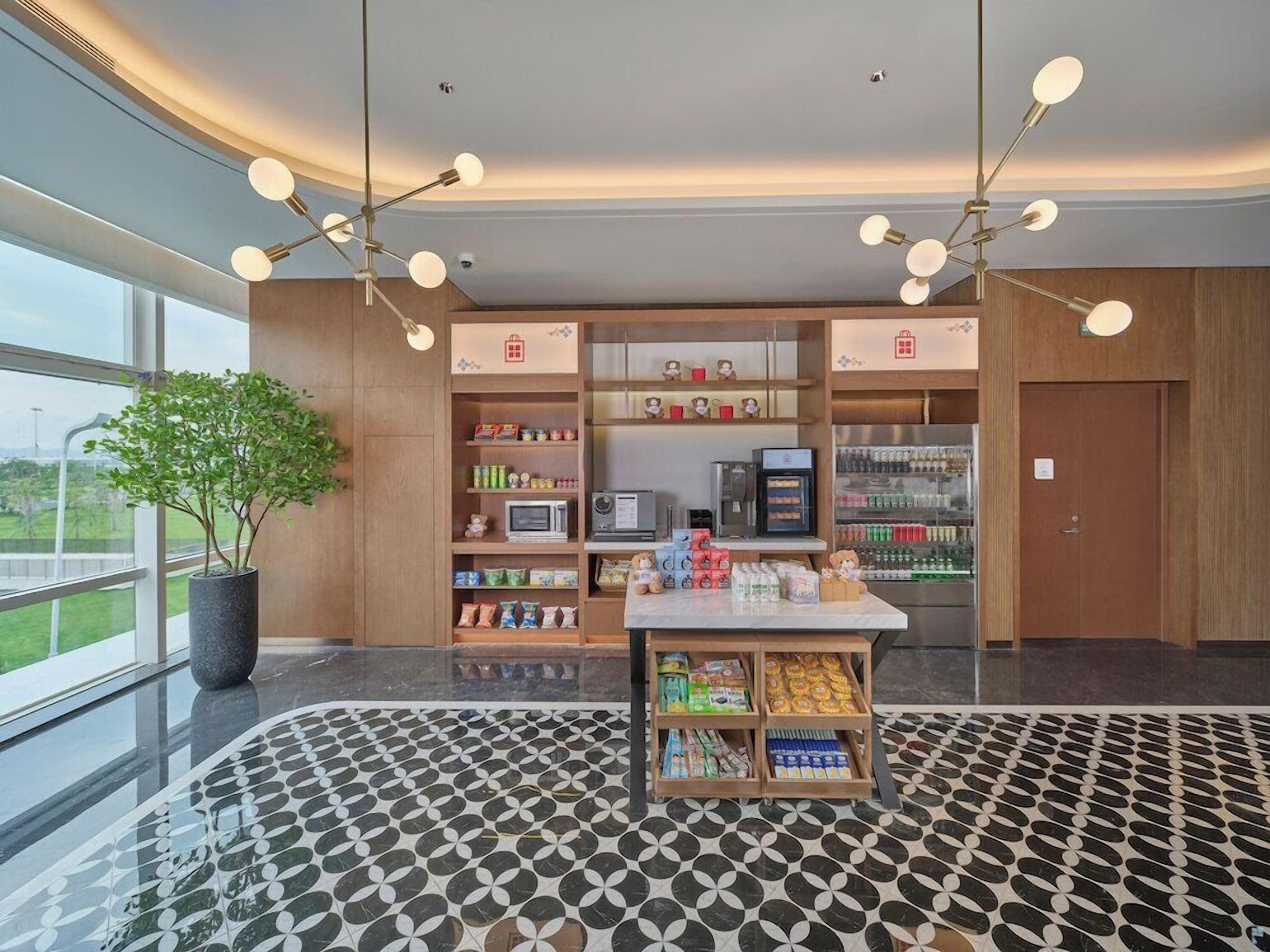 A modern hotel lobby with a central kiosk, wooden shelving with snacks, a microwave, and a potted plant.