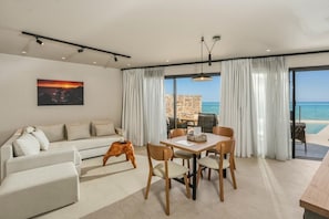 Un moderno salón con sofá, mesa de comedor, sillas, un cuadro en la pared y vista al mar a través de grandes ventanales.