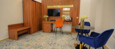 A room with a wooden desk, a television, a bench, and a dining area with blue chairs.