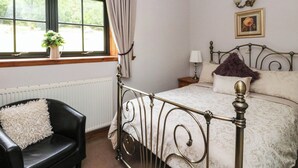 A bedroom with a bed, a chair, a window with curtains, a radiator, a nightstand, and a lamp. - Ardlui Church (Arrochar)
