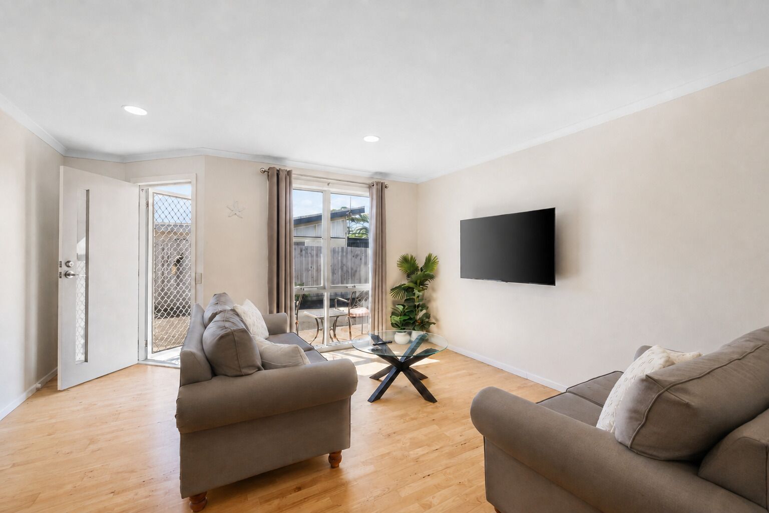 Una sala de estar con dos sofás, una mesa de centro de vidrio, un televisor de pantalla plana montado en la pared y una puerta corrediza de vidrio que da a un espacio exterior.