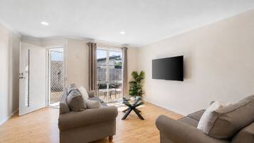 Una sala de estar con dos sofás, una mesa de centro de vidrio, un televisor de pantalla plana montado en la pared y una puerta corrediza de vidrio que da a un espacio exterior.