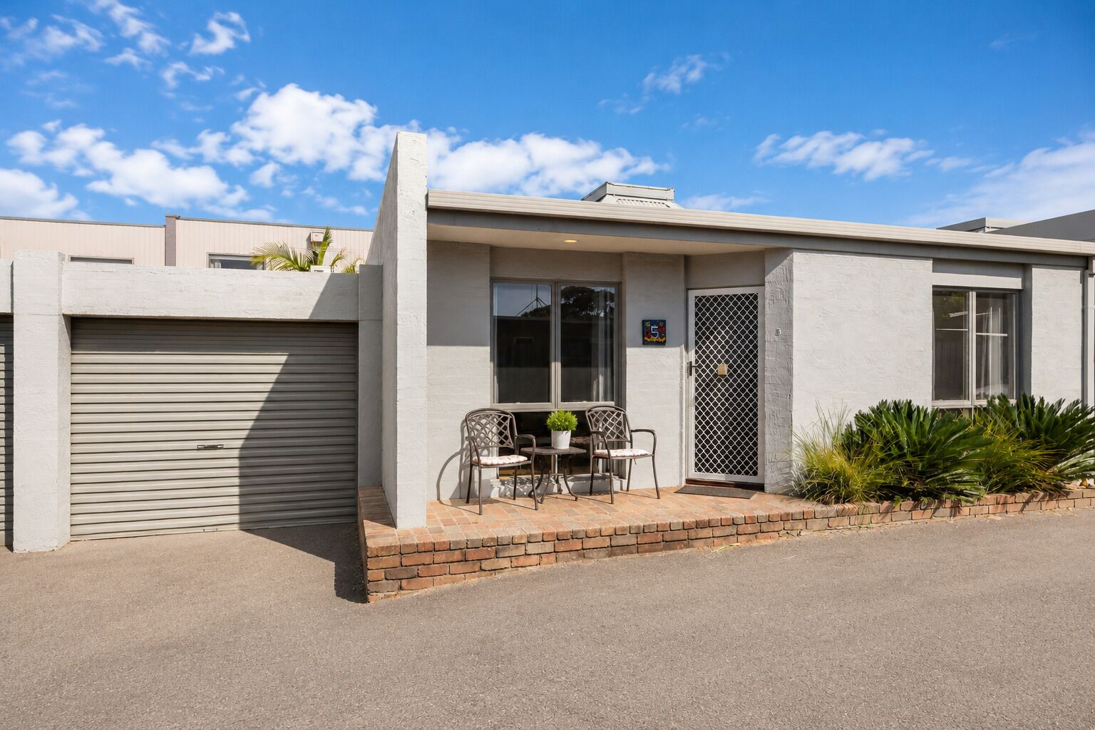 A modern building with a garage, a covered patio with chairs, and a small garden.