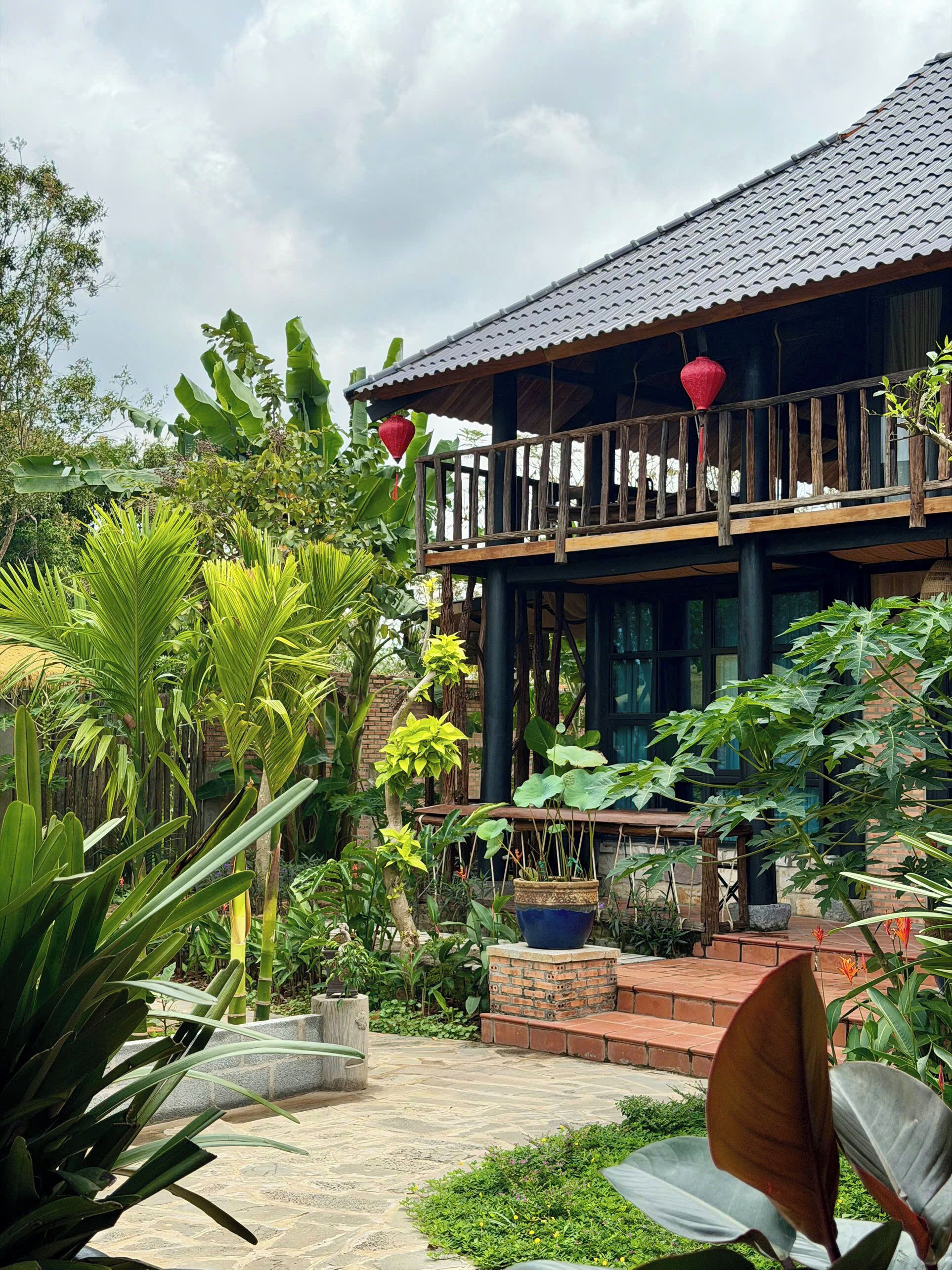A two-story building with a balcony, surrounded by lush greenery and a stone-paved courtyard.