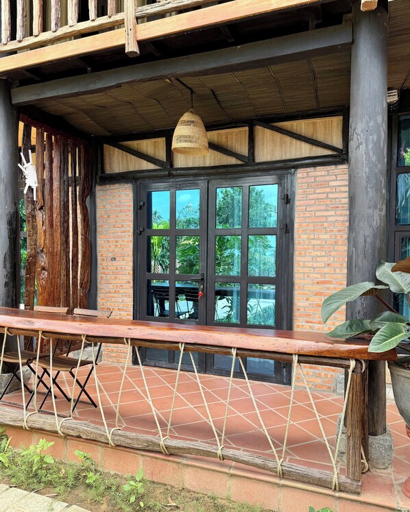 A rustic wooden balcony with a rope barrier, a brick wall, and a large window with a view of greenery. - Lam Mountain Retreat (Phu Quoc)