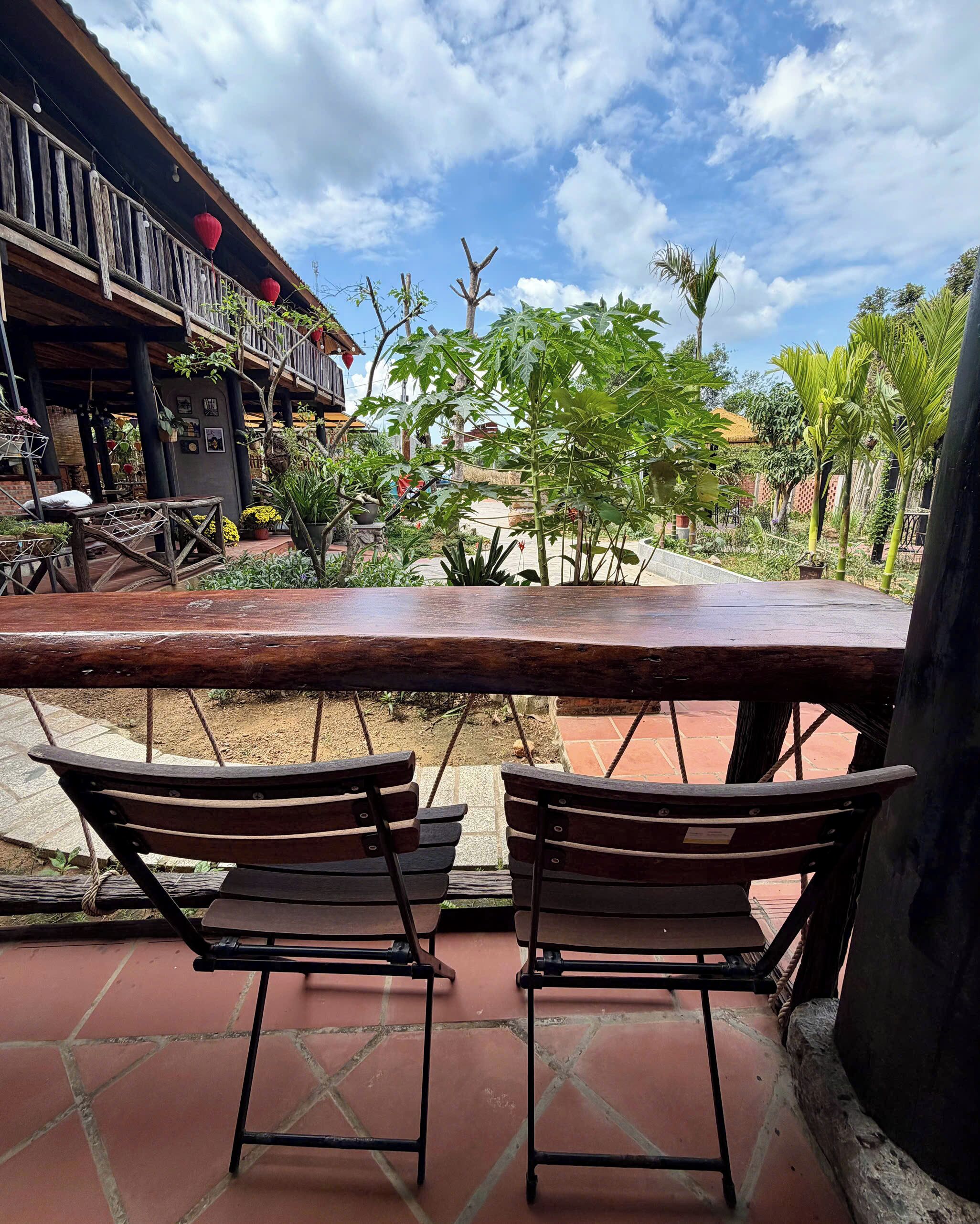An outdoor seating area with two wooden chairs and a bench, surrounded by potted plants and a building with a balcony.