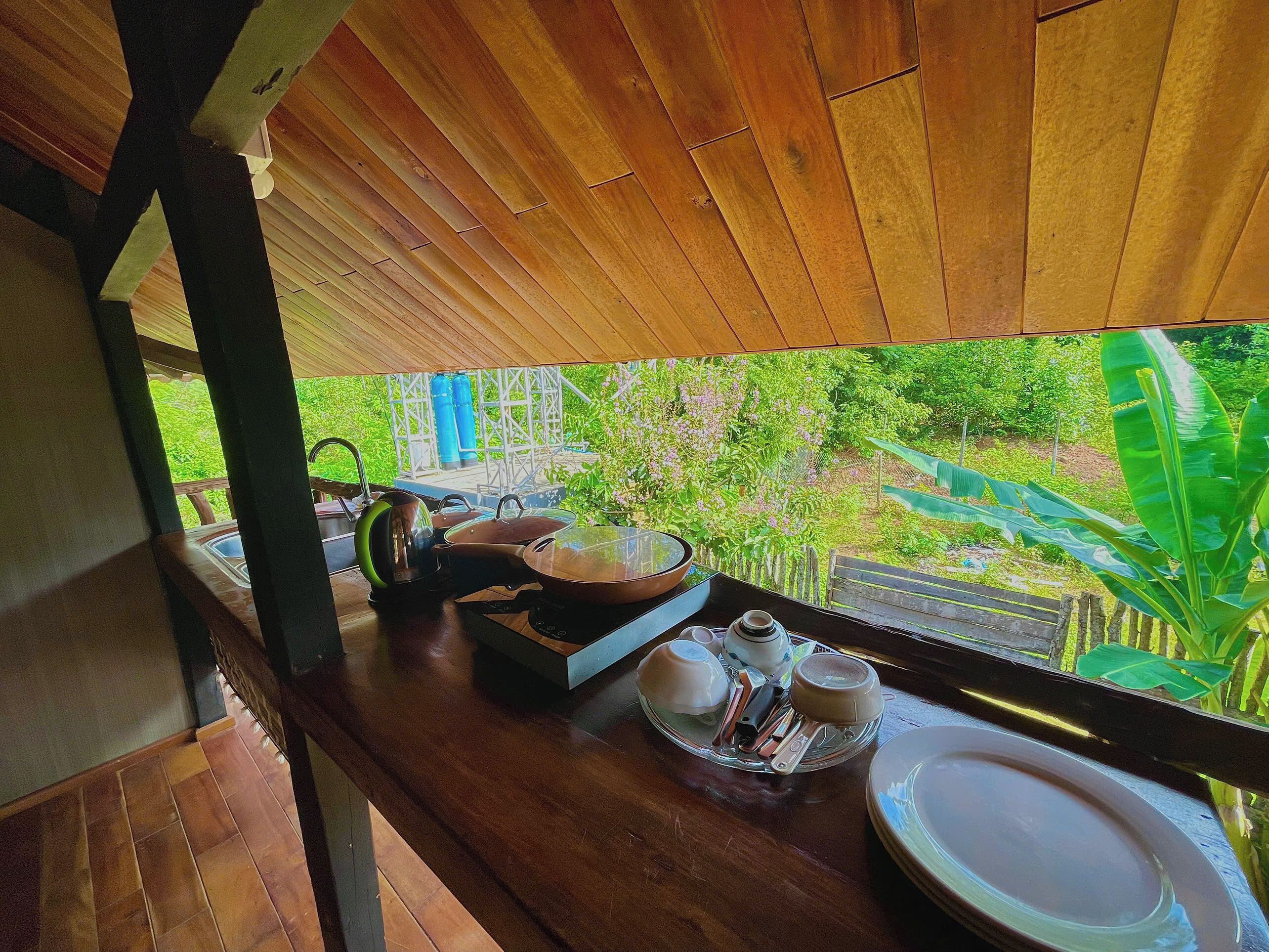 A wooden table with a view of greenery outside, a kettle, teapot, cups, and a tray with utensils.