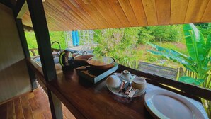 A wooden table with a view of greenery outside, a kettle, teapot, cups, and a tray with utensils.