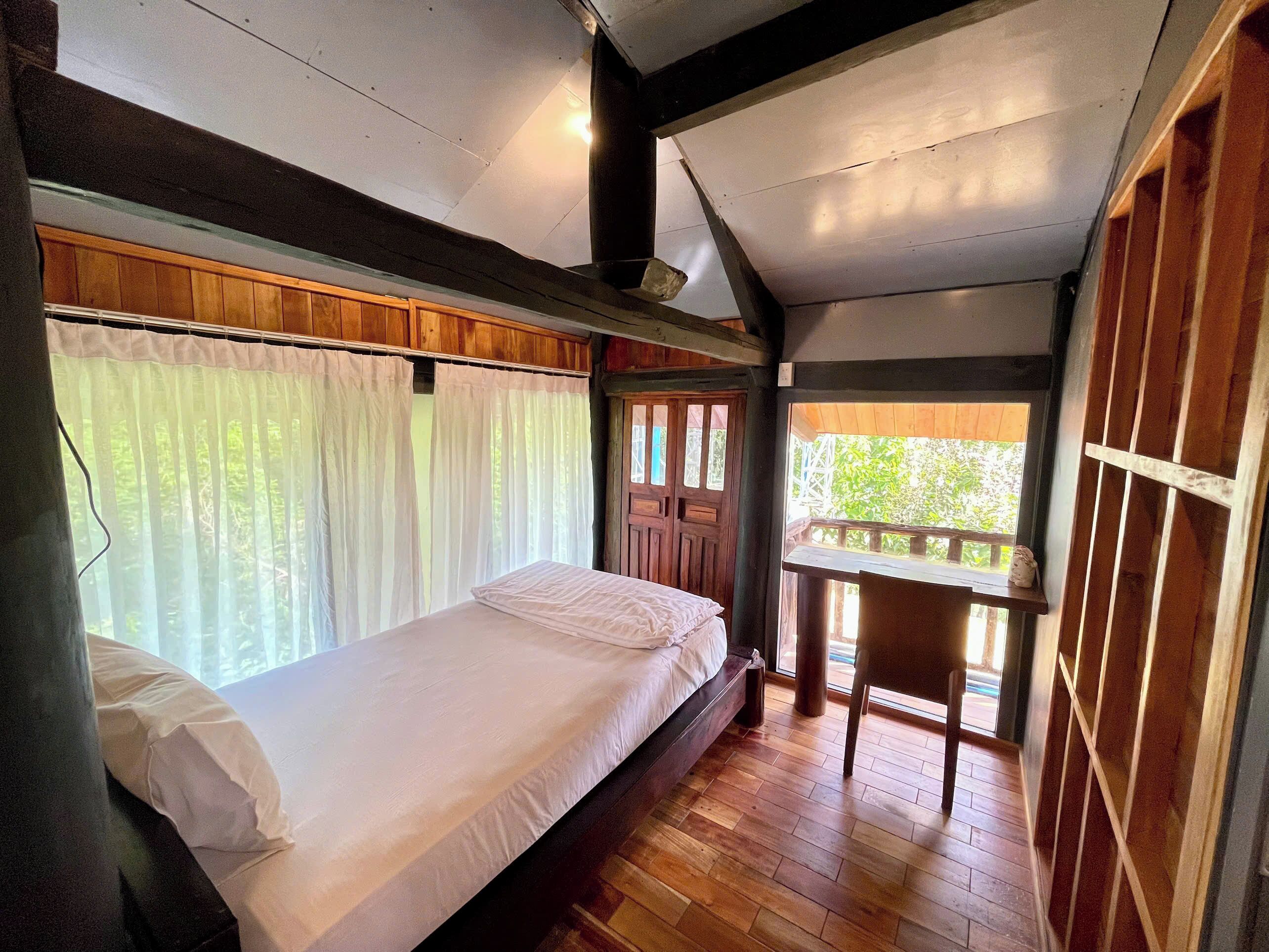 A single bed with a wooden headboard, a window with sheer curtains, and a view of greenery outside.