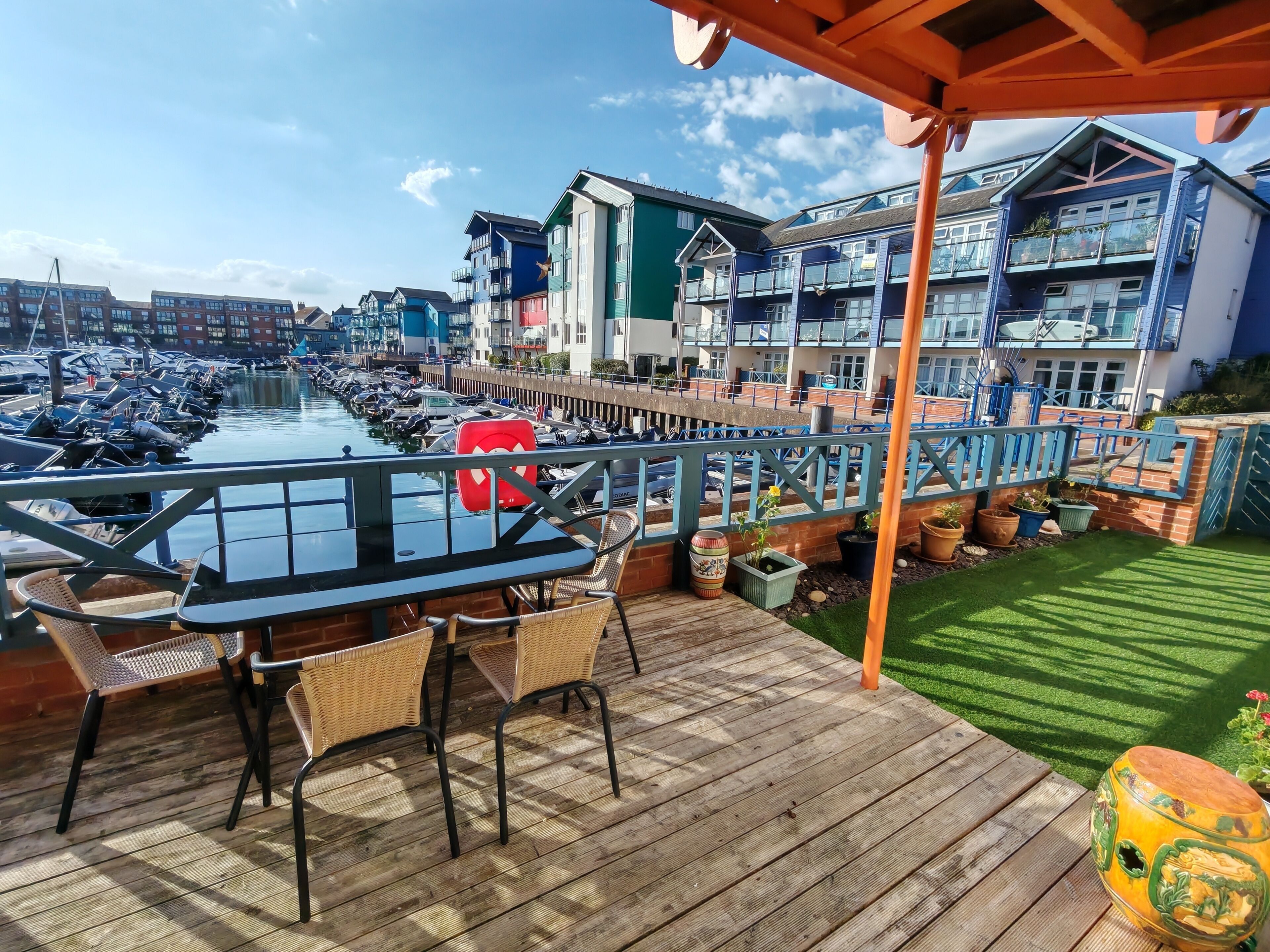 Une terrasse en bois avec une table et des chaises, donnant sur une marina où sont amarrés des bateaux, et sur une rangée de bâtiments aux couleurs vives.