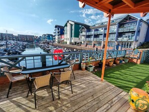 A wooden deck with a table and chairs, overlooking a marina with boats and a row of colorful buildings. - Waterfront Marina Apartment (Exmouth)