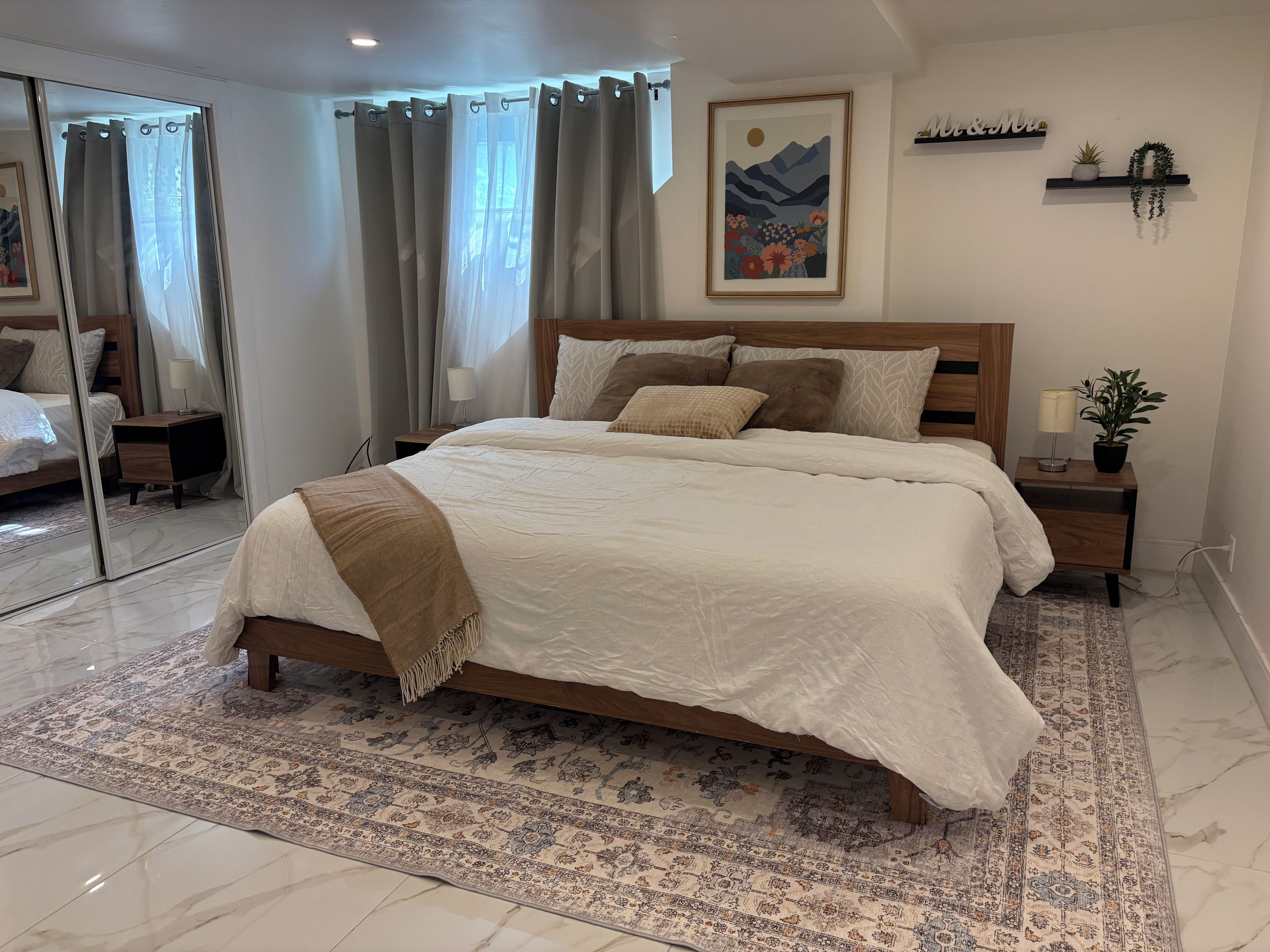 Ein Schlafzimmer mit einem großen Bett, einem Nachttisch, einem Spiegel, einem gerahmten Bild und einem Fenster mit Vorhängen.