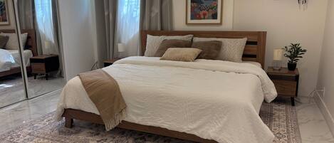 Un dormitorio con una cama grande, una mesita de noche, un espejo, un cuadro enmarcado y una ventana con cortinas.