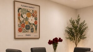 A dining area with a black table, four chairs, a vase with red flowers, and a framed artwork on the wall.