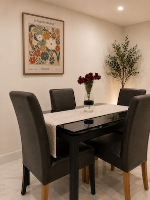 A dining area with a black table, four chairs, a vase with red flowers, and a framed artwork on the wall.