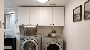 A laundry room with two front-loading washing machines, a basket on the left machine, and a shelf with jars and a plant on the right.