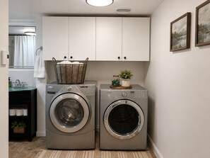 A laundry room with two front-loading washing machines, a basket on the left machine, and a shelf with jars and a plant on the right.