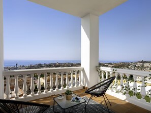 Un balcon donnant sur la mer, avec des balustrades blanches, deux chaises noires, une petite table agrémentée d’une plante et un magazine.