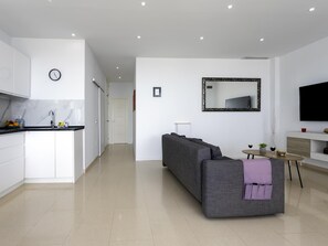 Ein moderner Wohnraum mit Küche, einem grauen Sofa, einem kleinen Tisch mit einer Vase sowie einem an der Wand befestigten Flachbildfernseher.