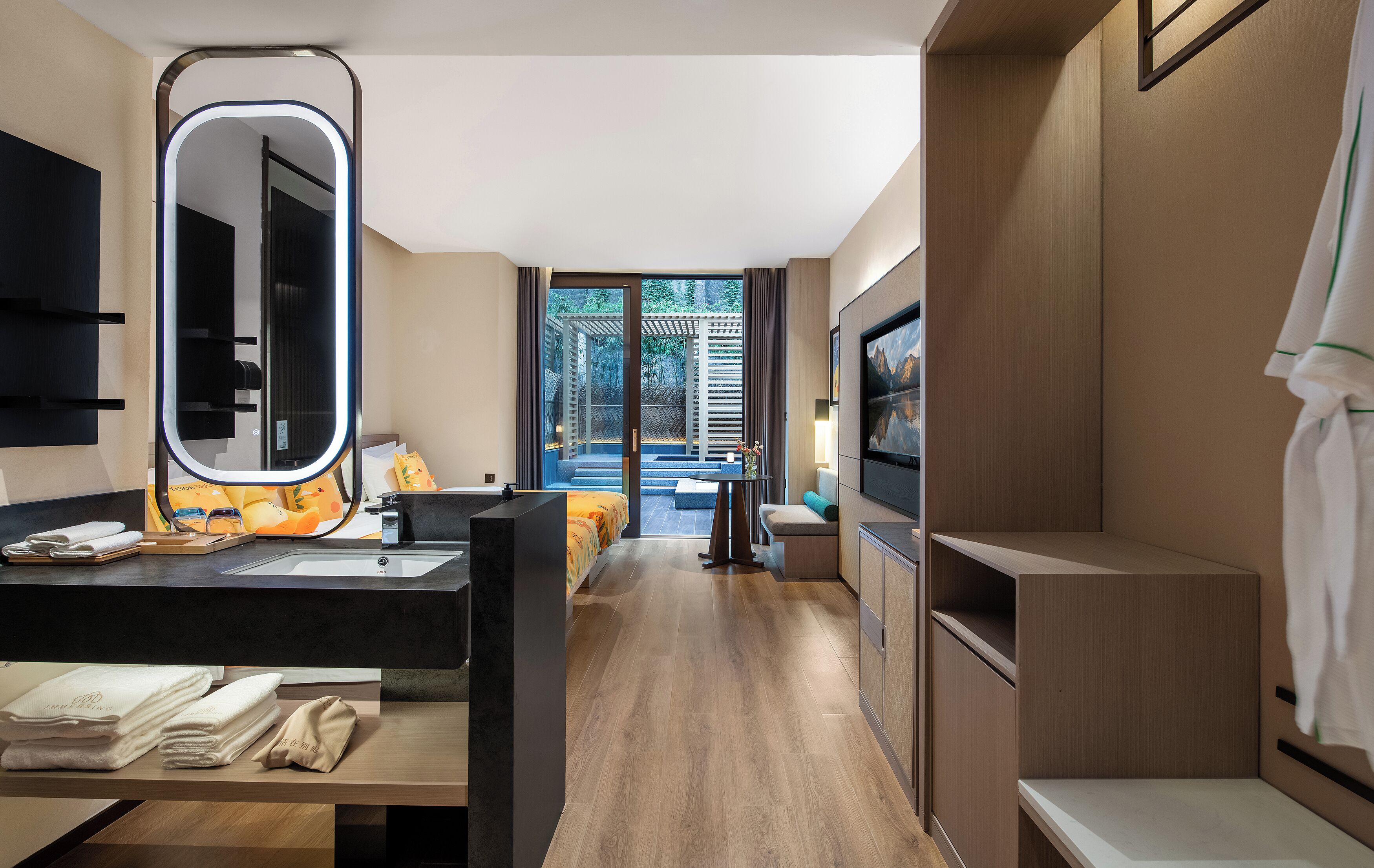 A modern hotel room with a large mirror, a bed with colorful bedding, a TV, and a view of a garden through the door.