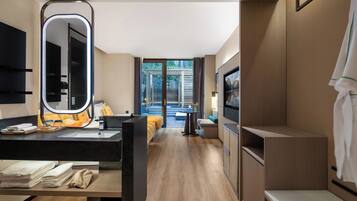 A modern hotel room with a large mirror, a bed with colorful bedding, a TV, and a view of a garden through the door.