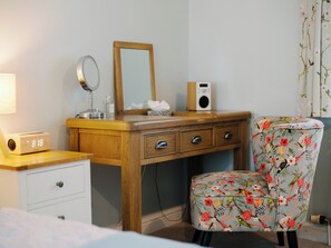 Ein Schlafzimmer mit einem hölzernen Schminktisch, einem Spiegel, einem Radio, einem blumigen Sessel und einem Bett.