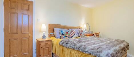 Un dormitorio con puerta de madera, una mesita de noche con lámpara, una cama con manta amarilla y varios almohadones, y una pequeña mesa con espejo.