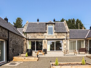 A stone building with a pitched roof, large windows, and a patio area with wooden benches and planters. - 5 bedroom accommodation in Auchtermuchty (Auchtermuchty)