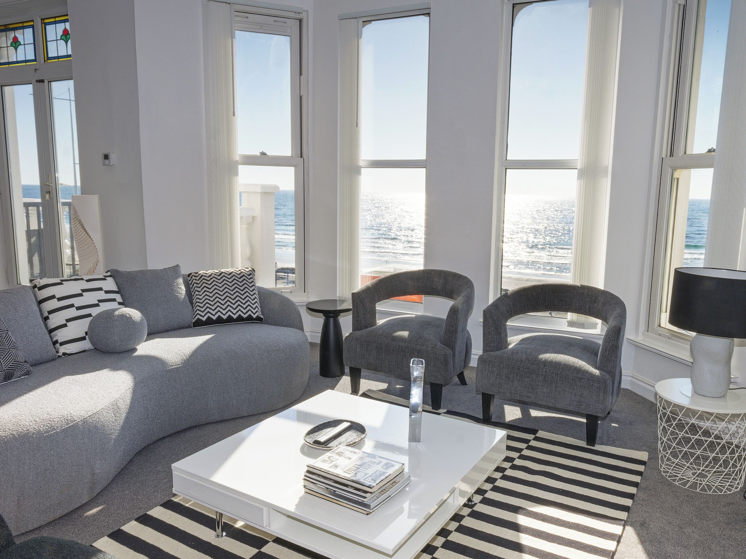 A modern living room with a sofa, armchairs, a coffee table, and large windows overlooking the sea.