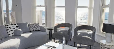 A modern living room with a sofa, armchairs, a coffee table, and large windows overlooking the sea.