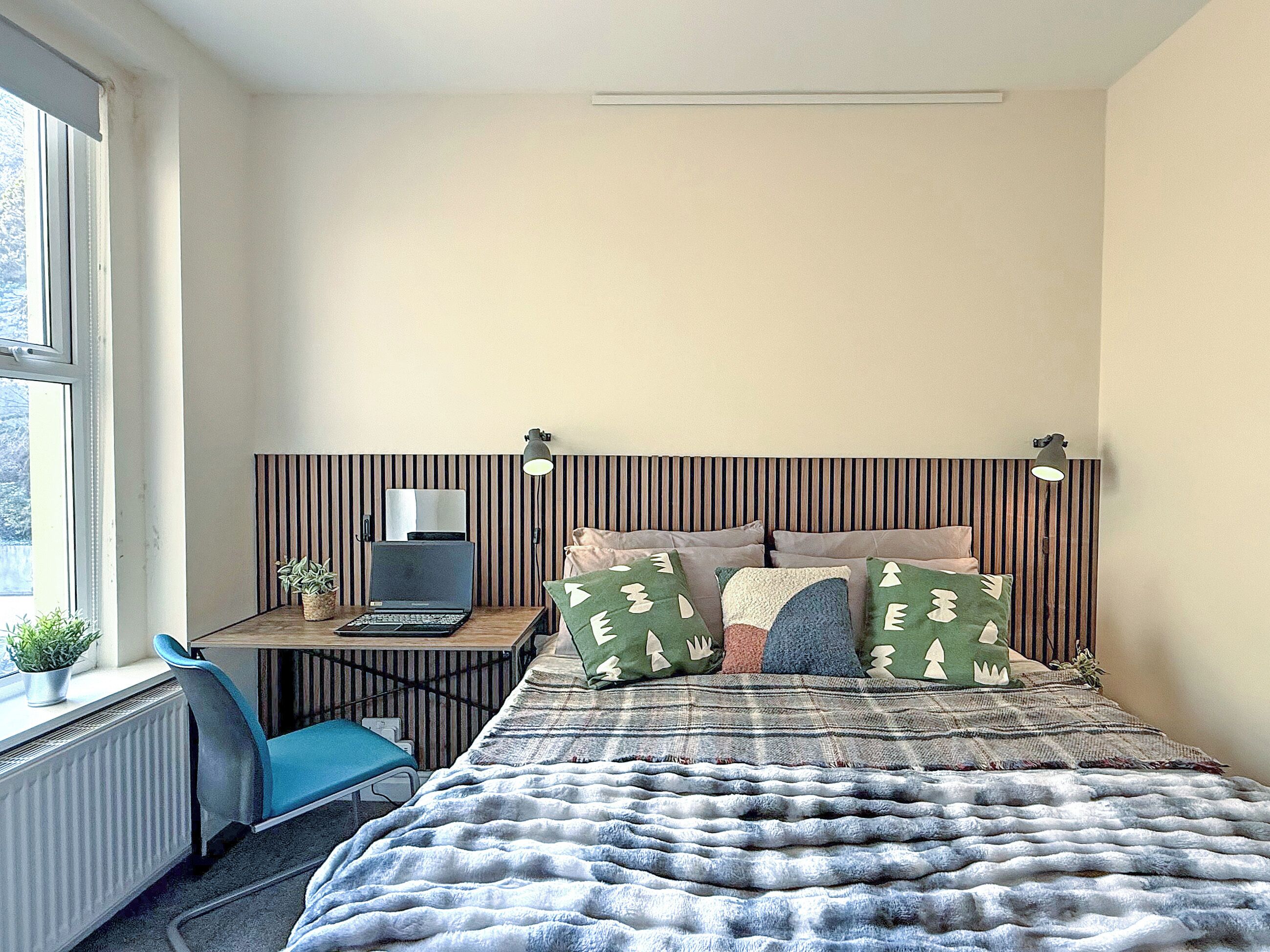 A bedroom with a bed, a desk with a laptop, a chair, a radiator, and a window.