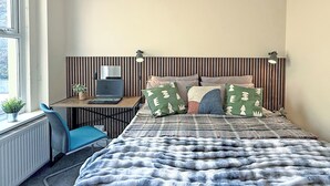 A bedroom with a bed, a desk with a laptop, a chair, a radiator, and a window.