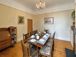 A dining room with a wooden table set for four, flanked by chairs with floral upholstery. A chandelier hangs from the ceiling, and there are framed pictures on the walls.