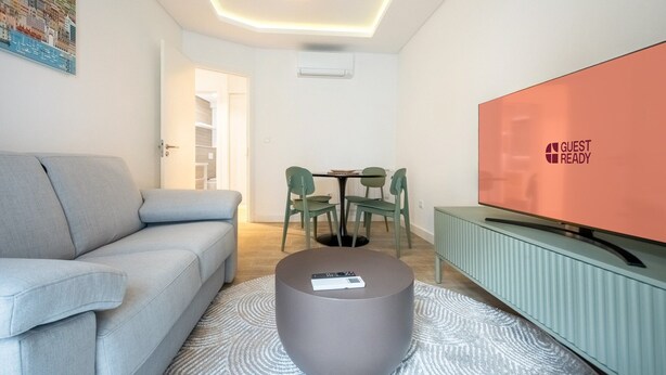 A modern living room with a grey sofa, a round coffee table, a flat-screen TV, and a dining area with a table and chairs. - GuestReady – Romantic Lisbon Nook (Lisbon)