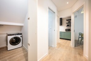 Une buanderie équipée d’un lave-linge, un couloir menant à une cuisine avec un four à micro-ondes et des placards, et un coin repas avec une chaise.