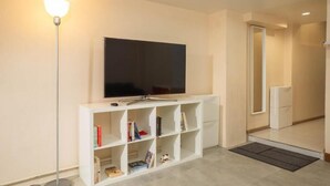 Ett modernt vardagsrum med en platt-TV som är monterad på en vit TV-bänk, en golvlampa och en bokhylla fylld med böcker.