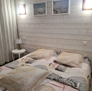 Ein Schlafzimmer mit einem Holzkopfteil, zwei Betten mit gemusterten Betttextilien, einem kleinen runden Tisch mit einer Lampe und zwei an der Wand befestigten Bilderrahmen.
