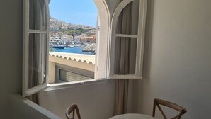 Quarto com vista para uma cidade costeira através de uma janela aberta com persianas brancas.